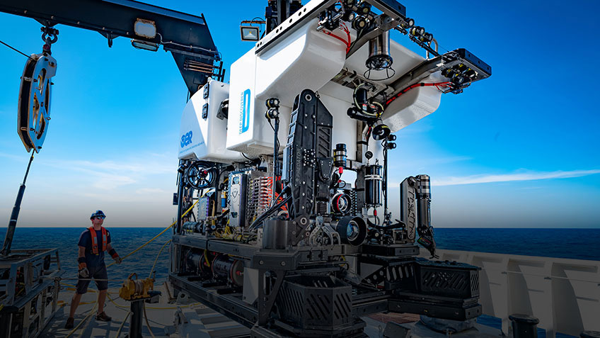 A remotely operated vehicle (ROV) on a ship deck with a person nearby, backed by ocean and sky.