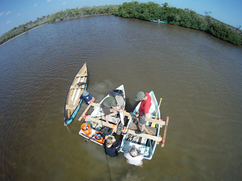 Collecting cores in the field required a team effort and coordination of equipment and people. The team makes use out of the floating coring platform and canoes to collect a core from the West Harbor area. Using a camera mounted on a 5-meter pole, Dominique Rissolo is able to capture an overhead view of the process.