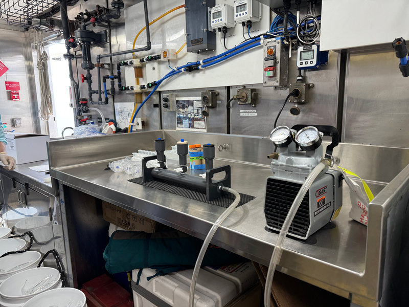 Image of the set up used to filter out the environmental DNA (eDNA) from collected water samples. With steady hands, we poured seawater from the 1- or 2-liter bags into the plastic cups which were attached to the three mini gray podiums. The boxy machine on the right is what causes the water to be sucked through the sample cup filters and it makes a loud sound while it's running.