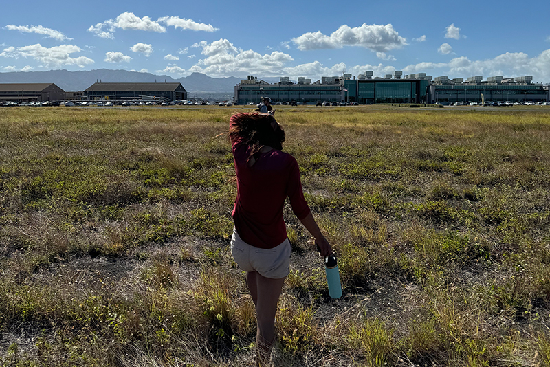 Image featuring explorer-in-training Claudia amongst others attempting a shortcut and walking through the prickly grass back to Okeanos Explorer. In the back, you can see the NOAA Daniel K. Inouye Regional Center.