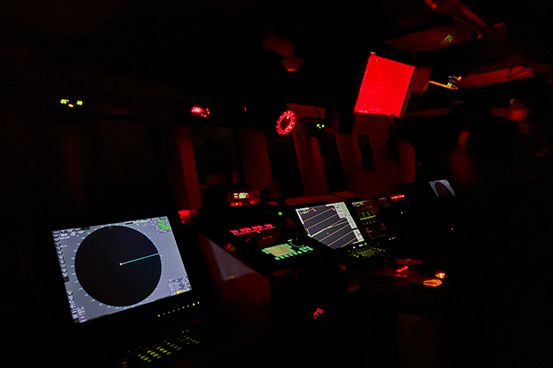 Image of the main controls within the bridge on NOAA Ship Okeanos Explorer. On the far left, you can see one of two radars used to detect surrounding things in the water. The radars can be turned up and down to reduce “noise” such as waves. At night, the bridge is kept dark with special lights used, which is why the screen on the top right has a red filter on it.