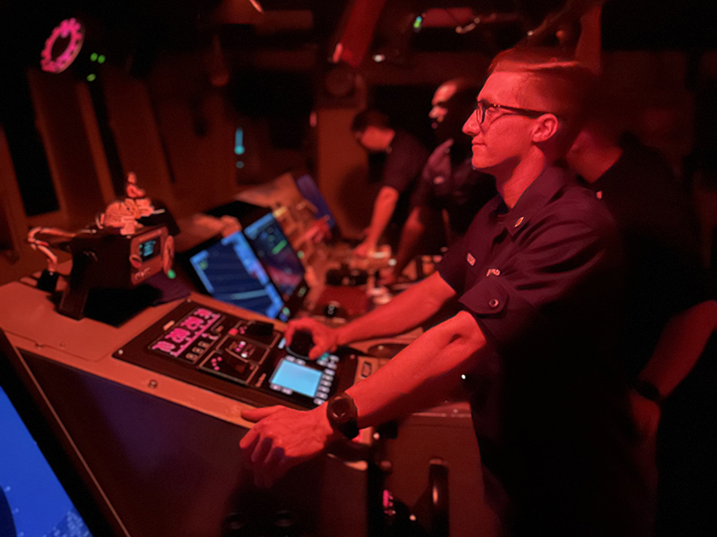 Person in uniform operating a control panel in a dimly lit, red-lit room.