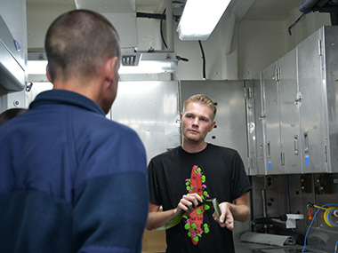 Makoa Pascoe, who is serving as a cultural liaison during the Beyond the Blue: Papahānaumokuākea Mapping 1 expedition, teaches members of NOAA Ship <i>Okeanos Explorer</i>’s crew how to make lei lā’ī of ti leaves. Prior to the ship’s departure, the cultural liaison team prepared a lei lāʻī for placement on the ship’s bridge rail as part of an Oli Pale to seek protection while underway. Many members of the on-ship team hung the lei they made on their stateroom doors.