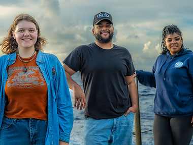 Image of three people standing on a vessel, with the sea and cloudy sky in the background.