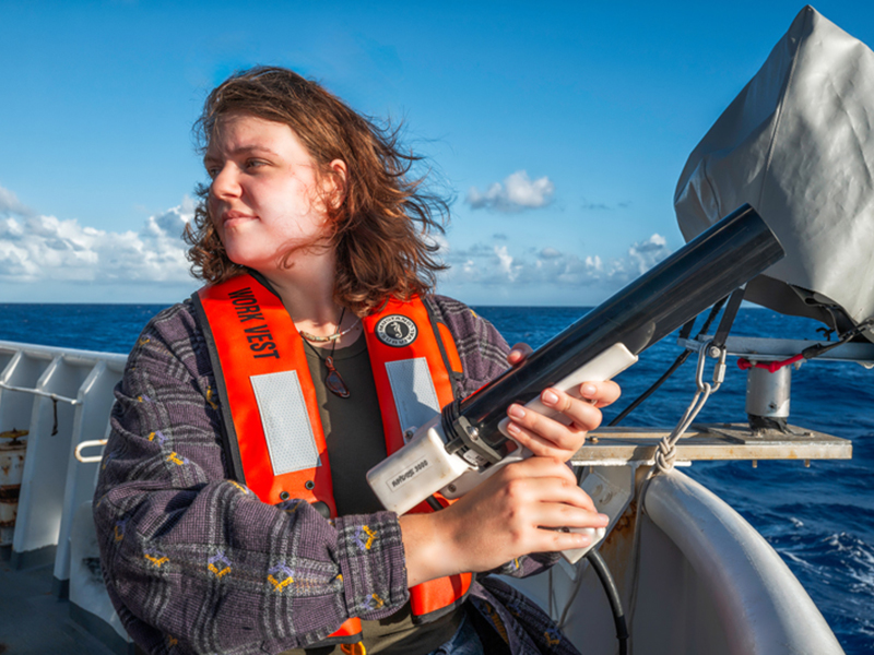 Person on a ship's deck holding a black cylindrical instrument, wearing an orange life vest, with the ocean and a clear sky in the background.