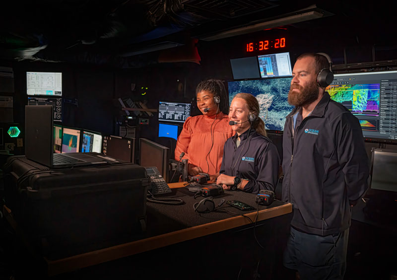 Three people in a control room with multiple monitors, wearing headsets.