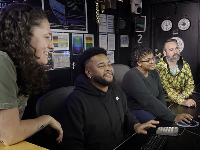 Four people in a control room working at computers, smiling and collaborating.