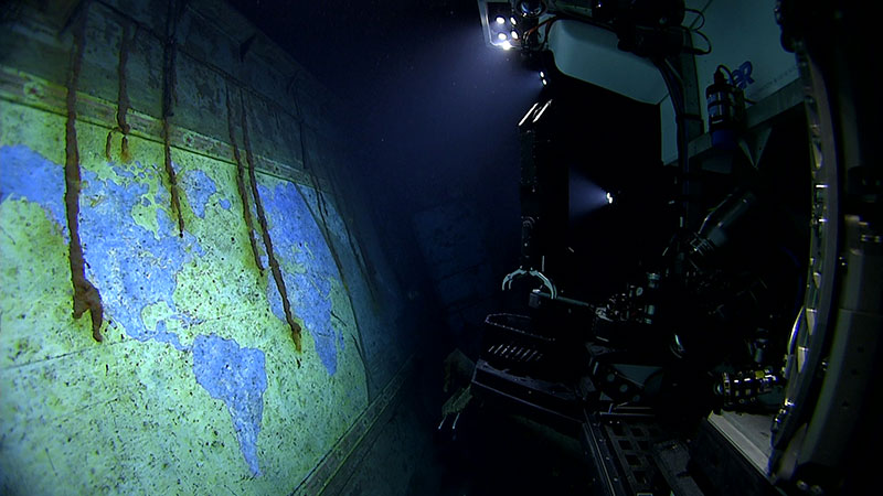Underwater scene with a robotic arm illuminating a faded world map on a shipwreck wall.