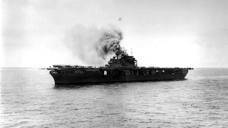 Aircraft carrier at sea emitting thick smoke under an overcast sky.