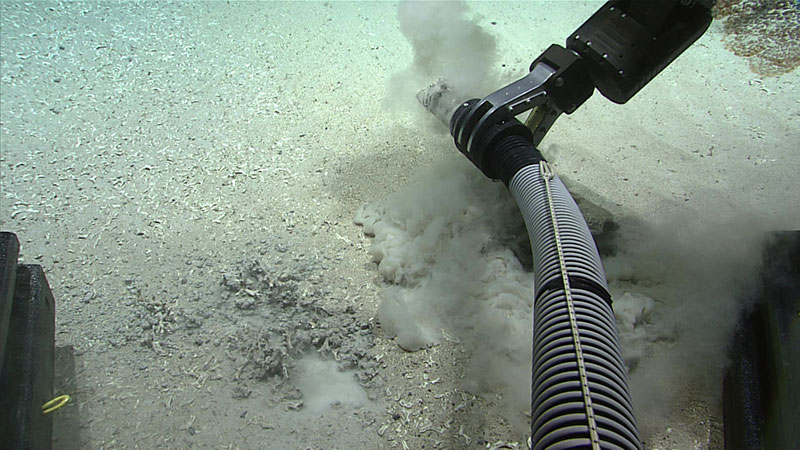 An underwater mechanical arm with a suction hose disturbing the ocean floor, creating sediment clouds.