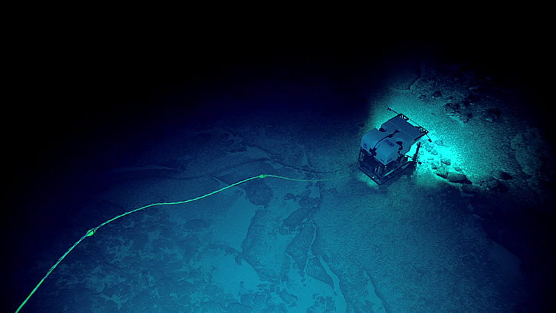 Underwater exploration vehicle with lights on, connected by a yellow cable on a dark seabed.