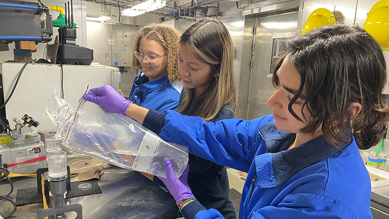 Three people in a lab, one pouring liquid from a plastic bag into a container, with lab equipment around.