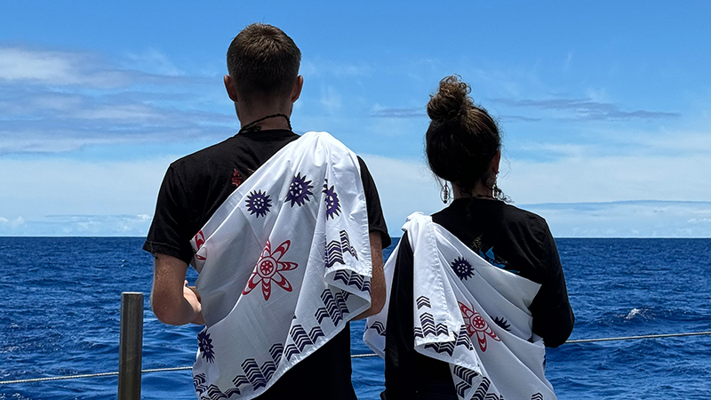 Two people on a boat looking at the ocean, wearing patterned white cloths over their shoulders.